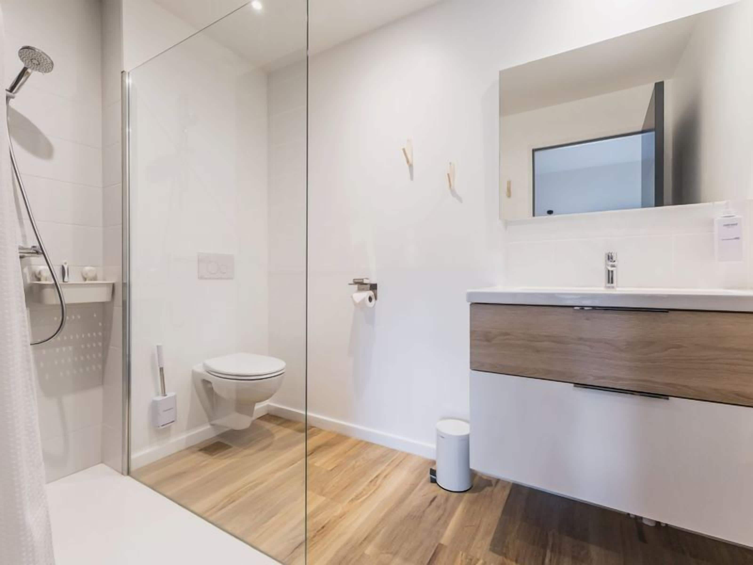 A modern bathroom with a glass shower enclosure, a toilet, a sink with a wooden vanity, and a mirror above the sink.