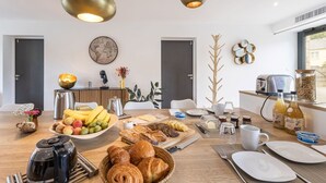 Une cuisine moderne avec une table en bois mise pour le petit-déjeuner, comprenant des fruits, des viennoiseries et du café.