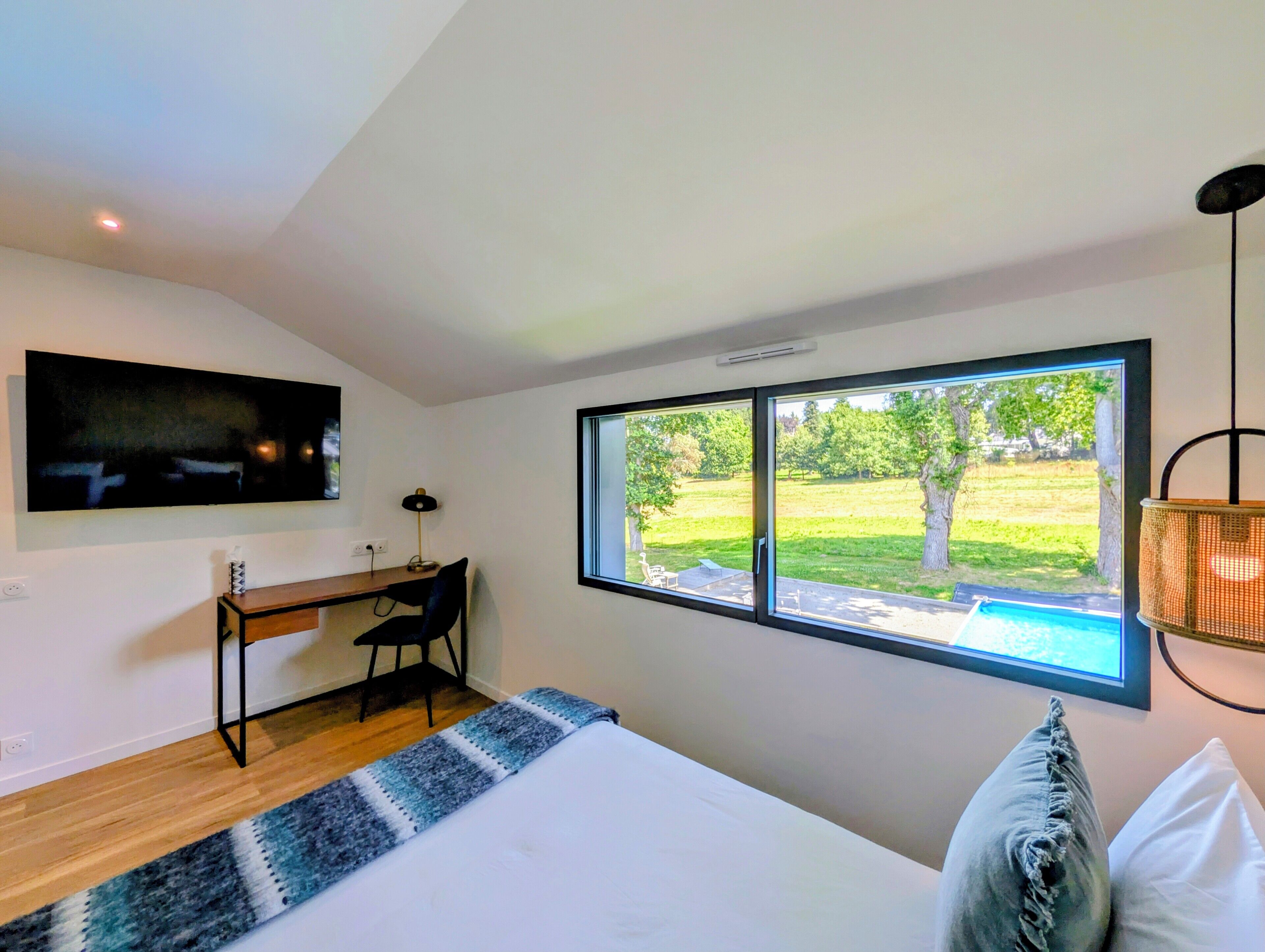 A bedroom with a bed, a desk, a chair, a TV, and a window with a view of a grassy area.