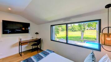 A bedroom with a bed, a desk, a chair, a TV, and a window with a view of a grassy area.
