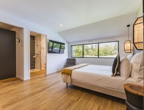 Een moderne slaapkamer met een groot bed, een houten vloer, een aan de muur bevestigde televisie en een raam met uitzicht op bomen.