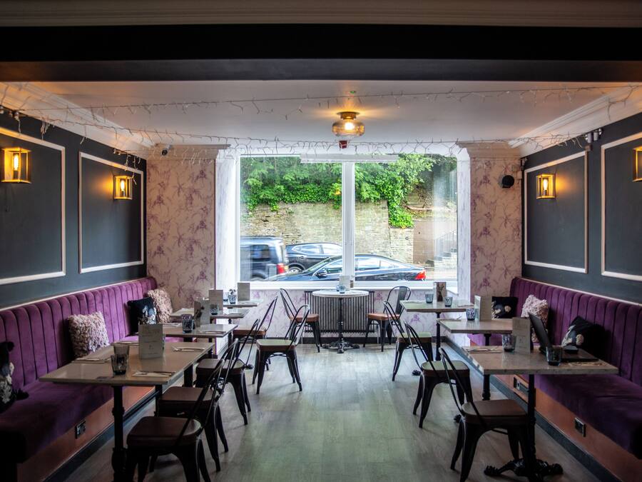 A restaurant with purple booths and wooden tables, a large window with a view of parked cars, and decorative string lights.