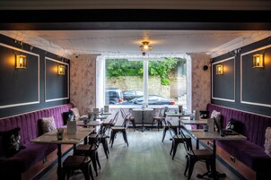 A restaurant with purple booths and wooden tables, a large window with a view of parked cars, and decorative string lights.
