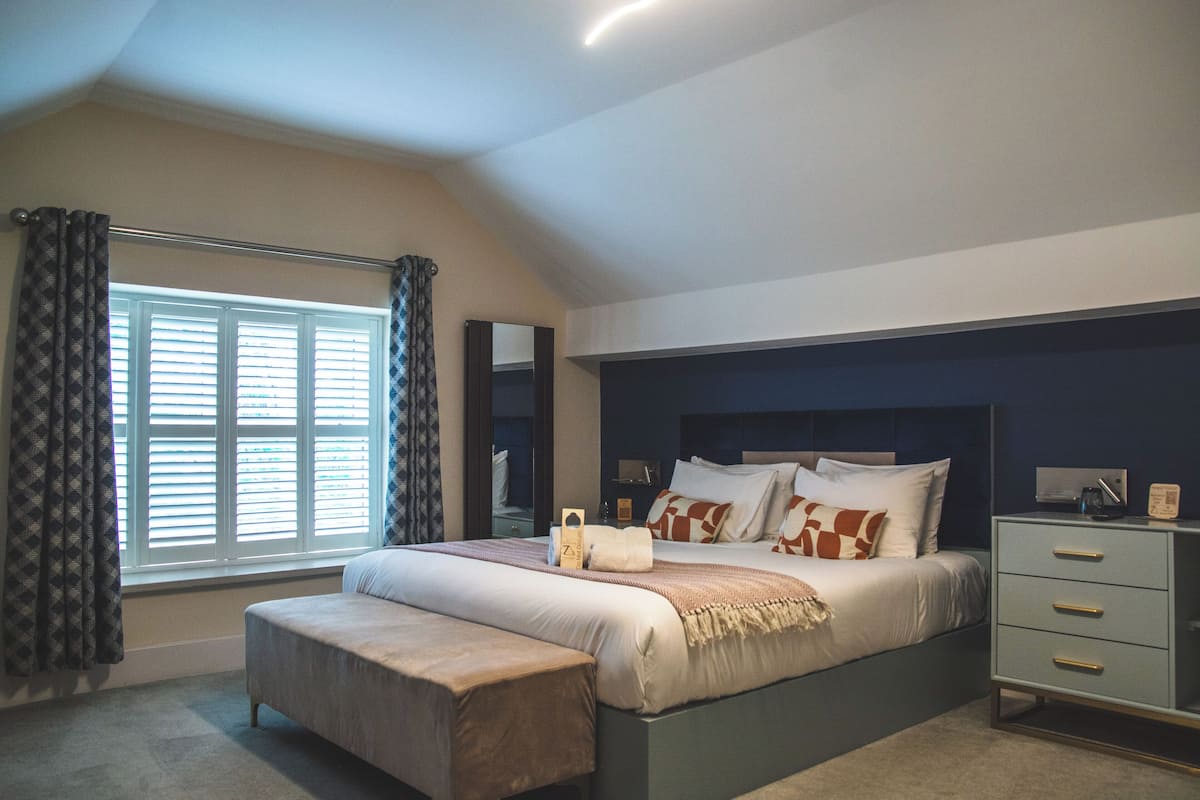 Une chambre à coucher avec un lit, une banquette, une table de chevet et une fenêtre avec des stores.