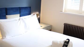 Una cama bien tendida con sábanas blancas, un cabecero azul y una ventana con persianas.