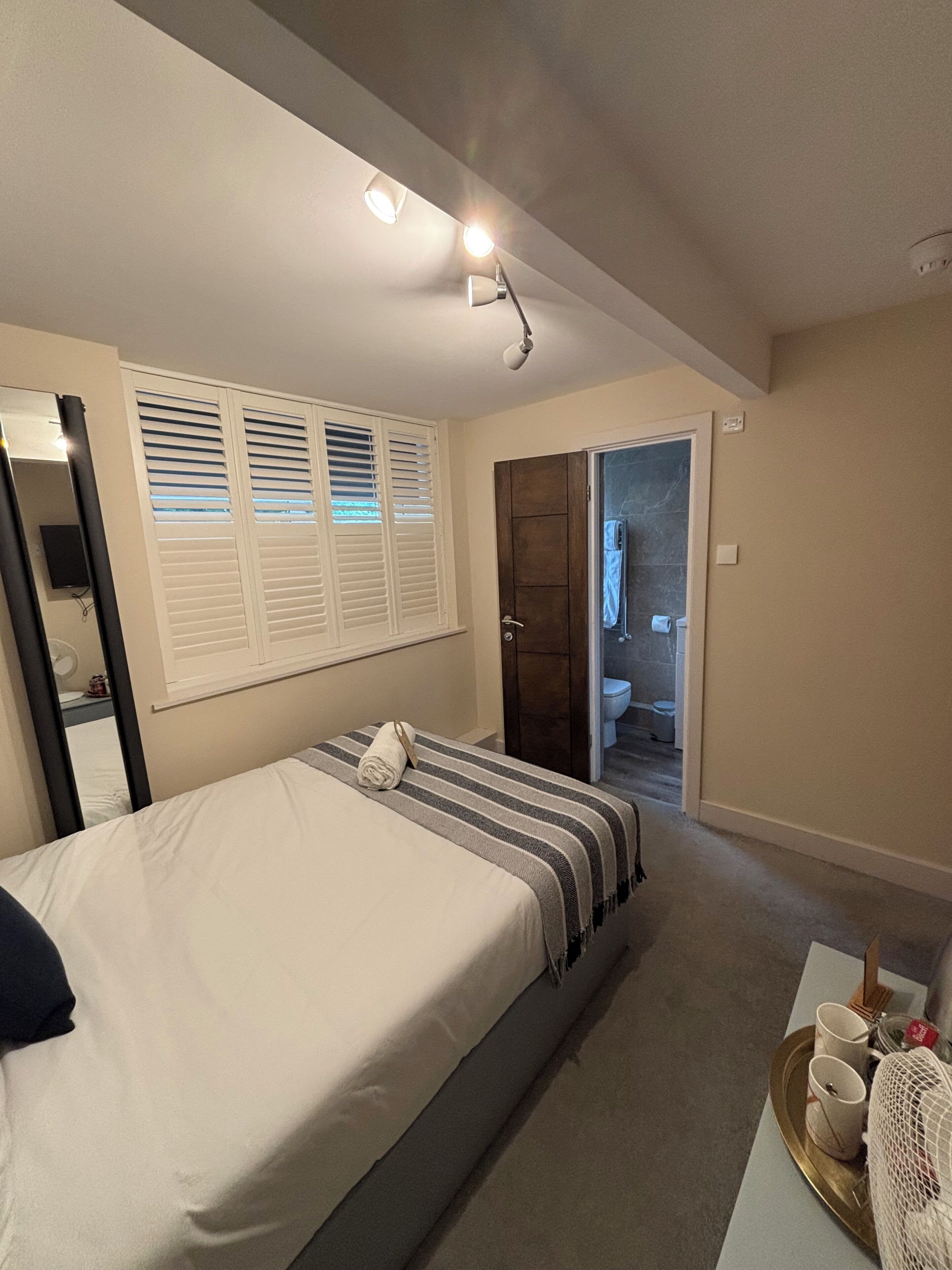 Een slaapkamer met een bed, een nachtkastje met kopjes, een raam met zonwering en een deur naar een badkamer.