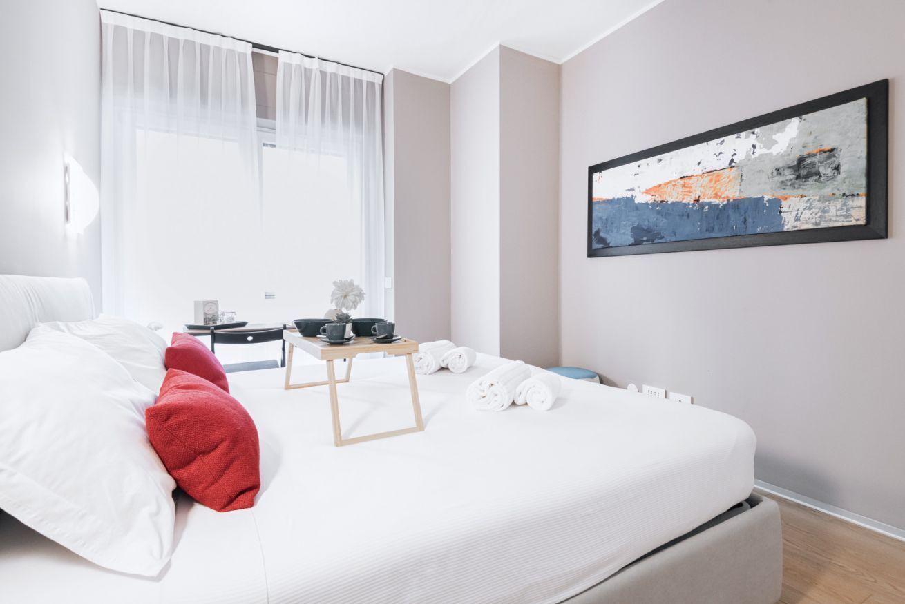 A modern hotel room with a neatly made bed, a bedside table with a lamp, a small table with cups and a plant, and a large abstract painting on the wall.