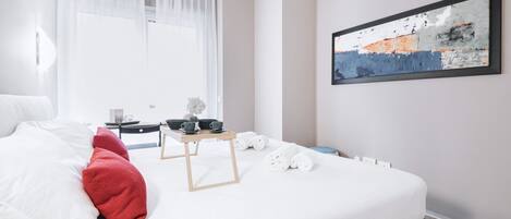 A modern hotel room with a neatly made bed, a bedside table with a lamp, a small table with cups and a plant, and a large abstract painting on the wall.
