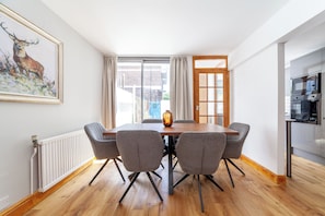 A dining area with a wooden table and grey chairs, a radiator, a painting of a deer, and a kitchen with modern appliances.