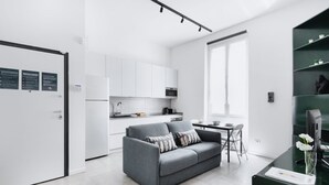 Una cocina moderna con armarios blancos, un sofá gris con cojines estampados y un comedor con una mesa pequeña y sillas.
