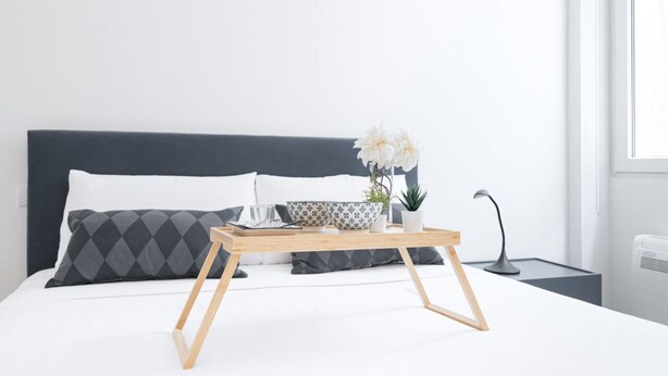 Una cama bien hecha con un cabecero oscuro, una mesita de noche con una lámpara y una ventana con luz natural.