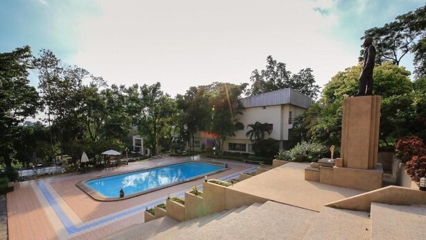 A swimming pool surrounded by a landscaped garden with a statue, a building in the background, and a clear sky. - Rajpracha Pakchong (Pak Chong)