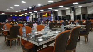 A spacious dining area with multiple tables and chairs, water bottles on tables, and a stage with a large screen in the background. - Celik Hanim Termal Otel & Spa (Ulukisla)