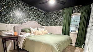 Ein Schlafzimmer mit floralem Wandpapier, einem Bett mit grüner Bettdecke, einem hölzernen Nachttisch und einem Deckenventilator.