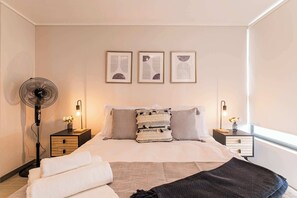 Un dormitorio con cama, mesitas de noche, ventilador de pie, decoración en la pared y una ventana.