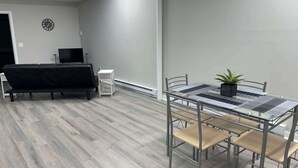 A modern room with a glass table, metal chairs, a black sofa, a small white side table, a clock on the wall, and a potted plant. - Private Apartment in Southbury 102 (Southbury)