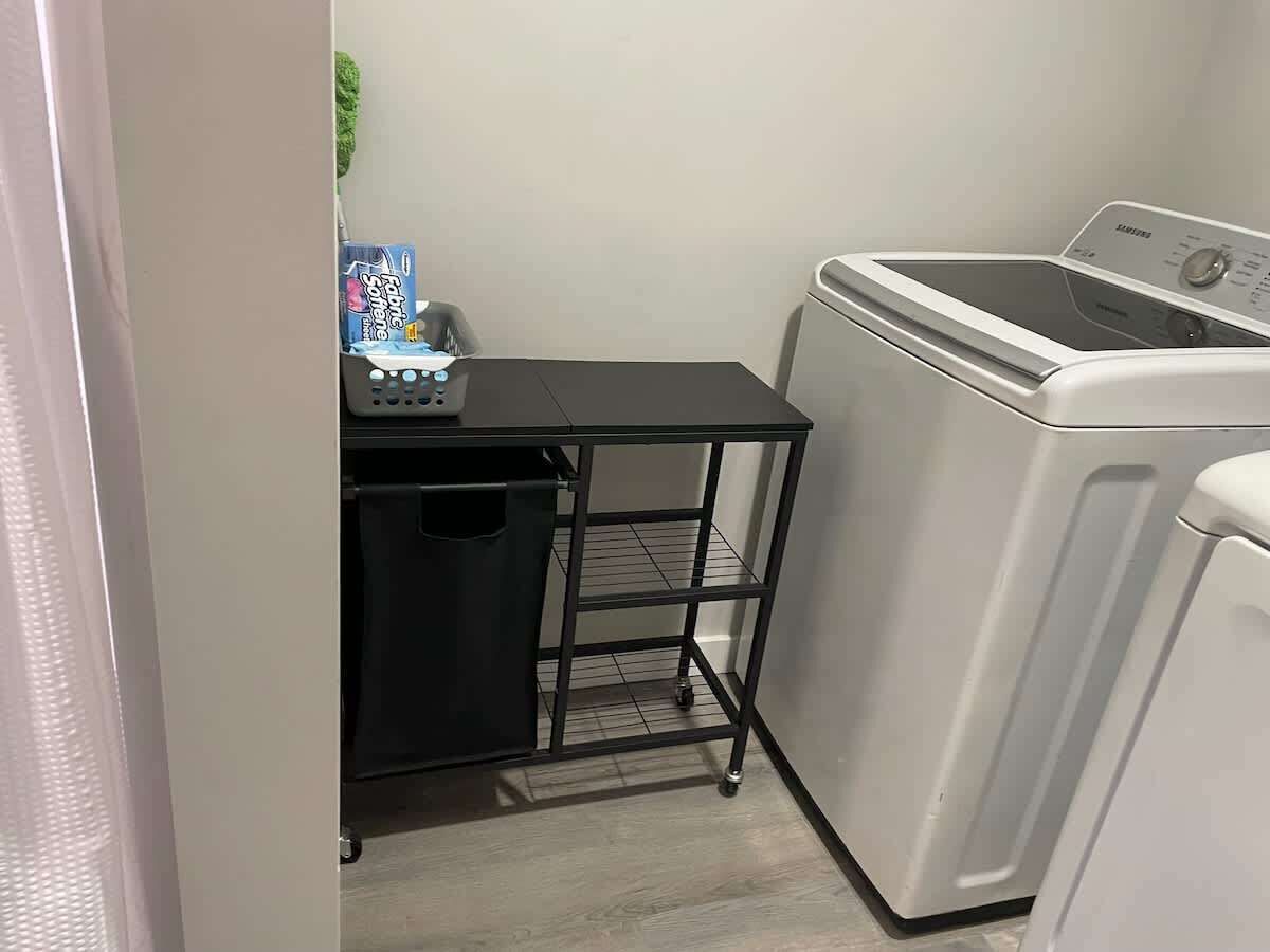 A laundry area with a washing machine, a small black metal cart with a basket, and a trash bin.