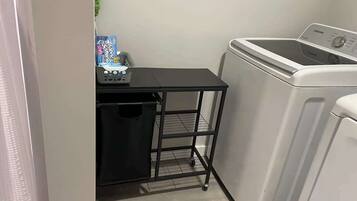 A laundry area with a washing machine, a small black metal cart with a basket, and a trash bin.