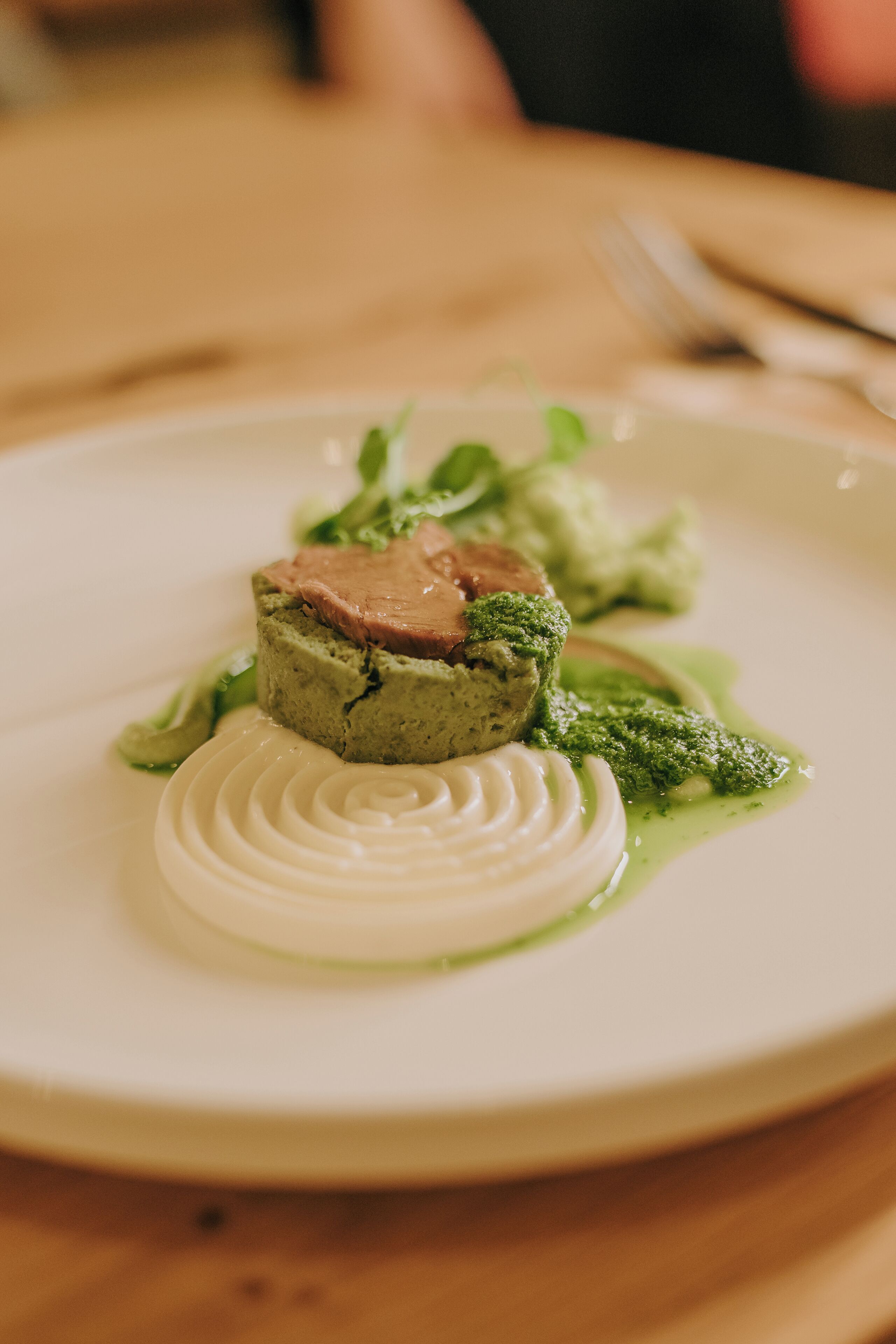 A white plate with a layered dish, including a green base, a white swirl, and a pink topping, accompanied by green sauce and greens.