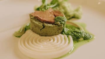 A white plate with a layered dish, including a green base, a white swirl, and a pink topping, accompanied by green sauce and greens.