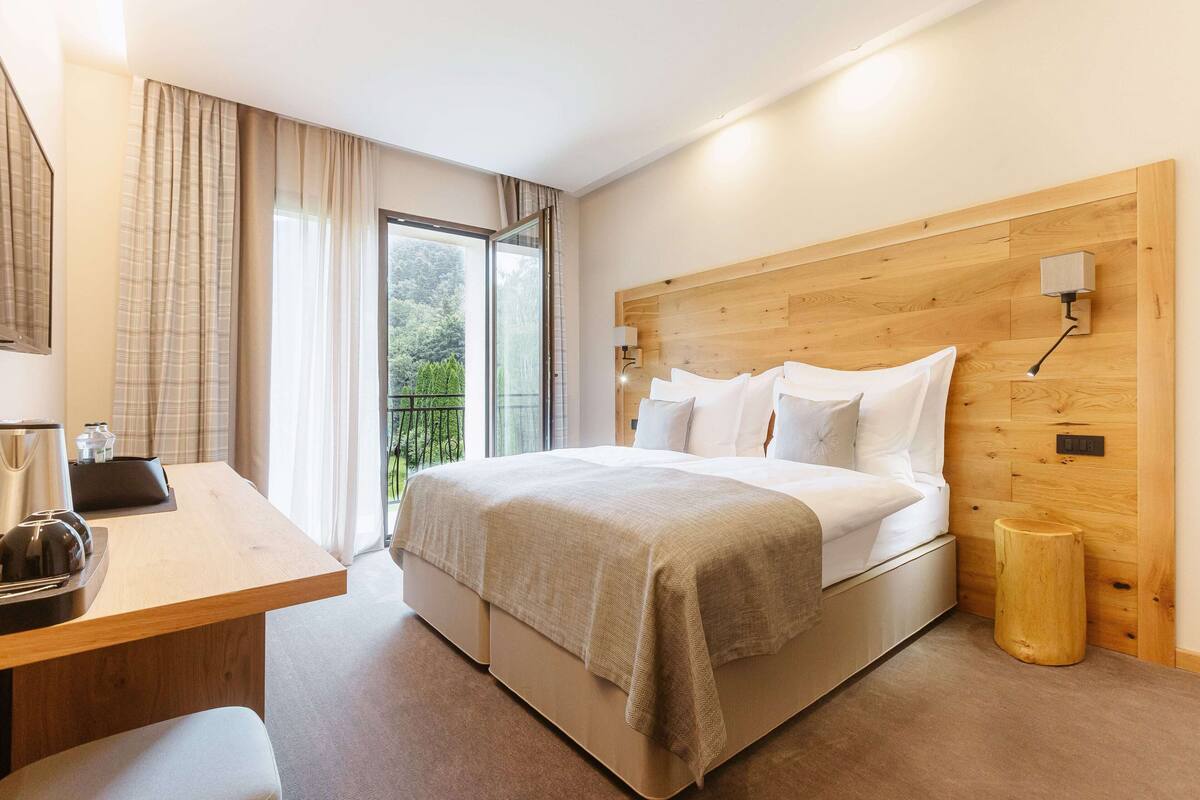 A modern hotel room with a wooden headboard, a large bed with white linens, a bedside table with a kettle, and a view of greenery through the window.