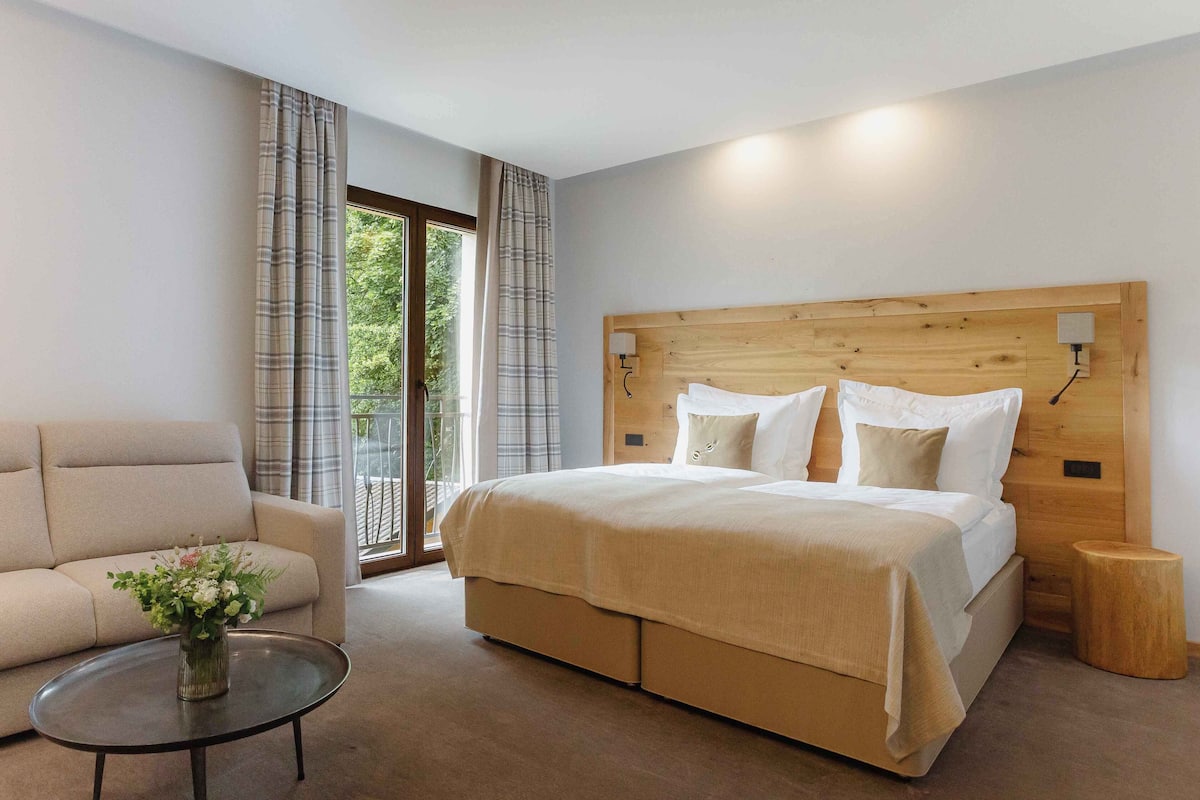 A modern bedroom with a large bed, a sofa, a small table, and a view of greenery outside.