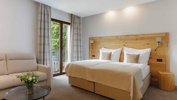 A modern bedroom with a large bed, a sofa, a small table, and a view of greenery outside.