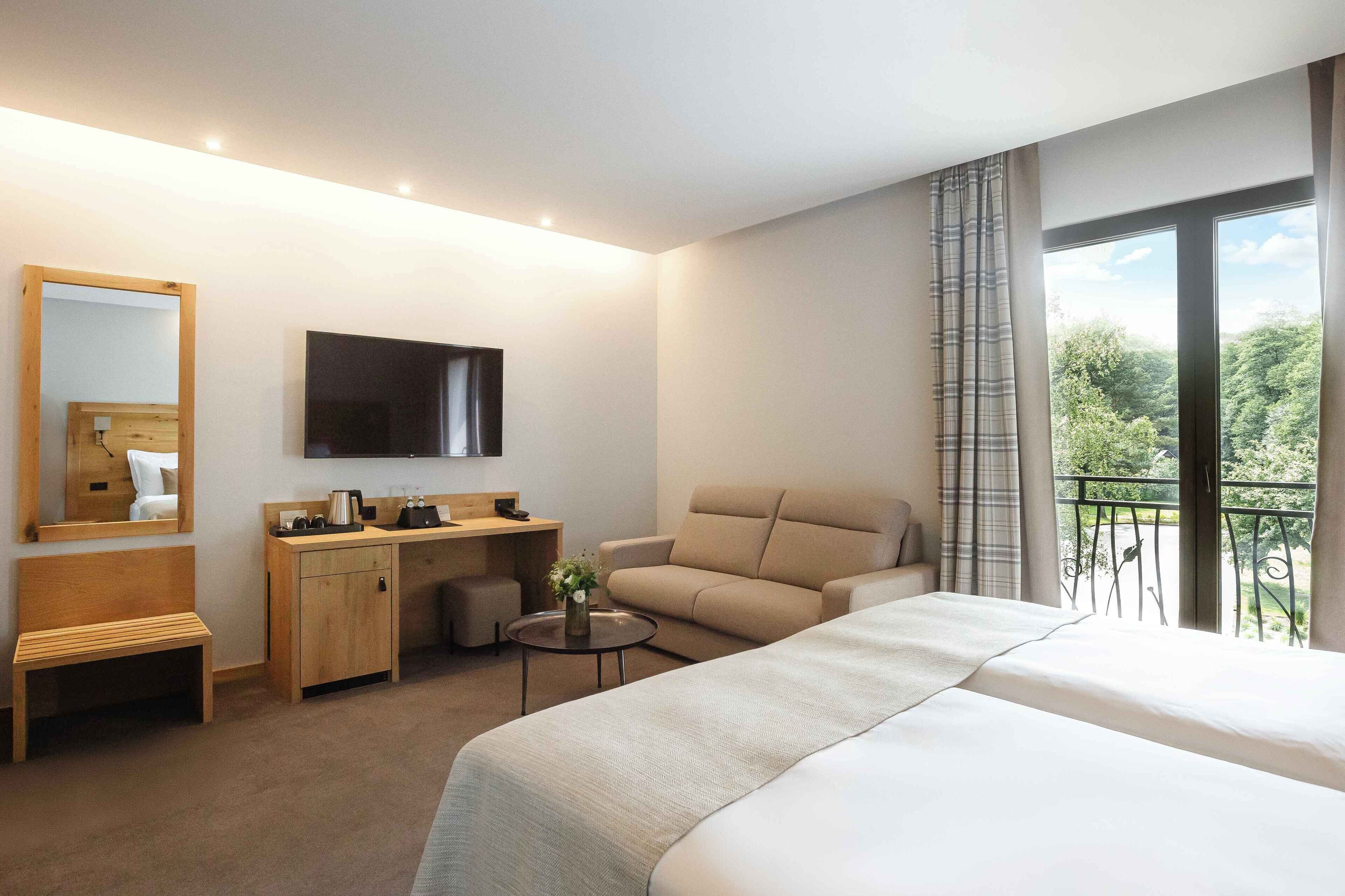 A hotel room with a bed, a sofa, a desk with a chair, a small table with a vase, a mirror, and a TV.