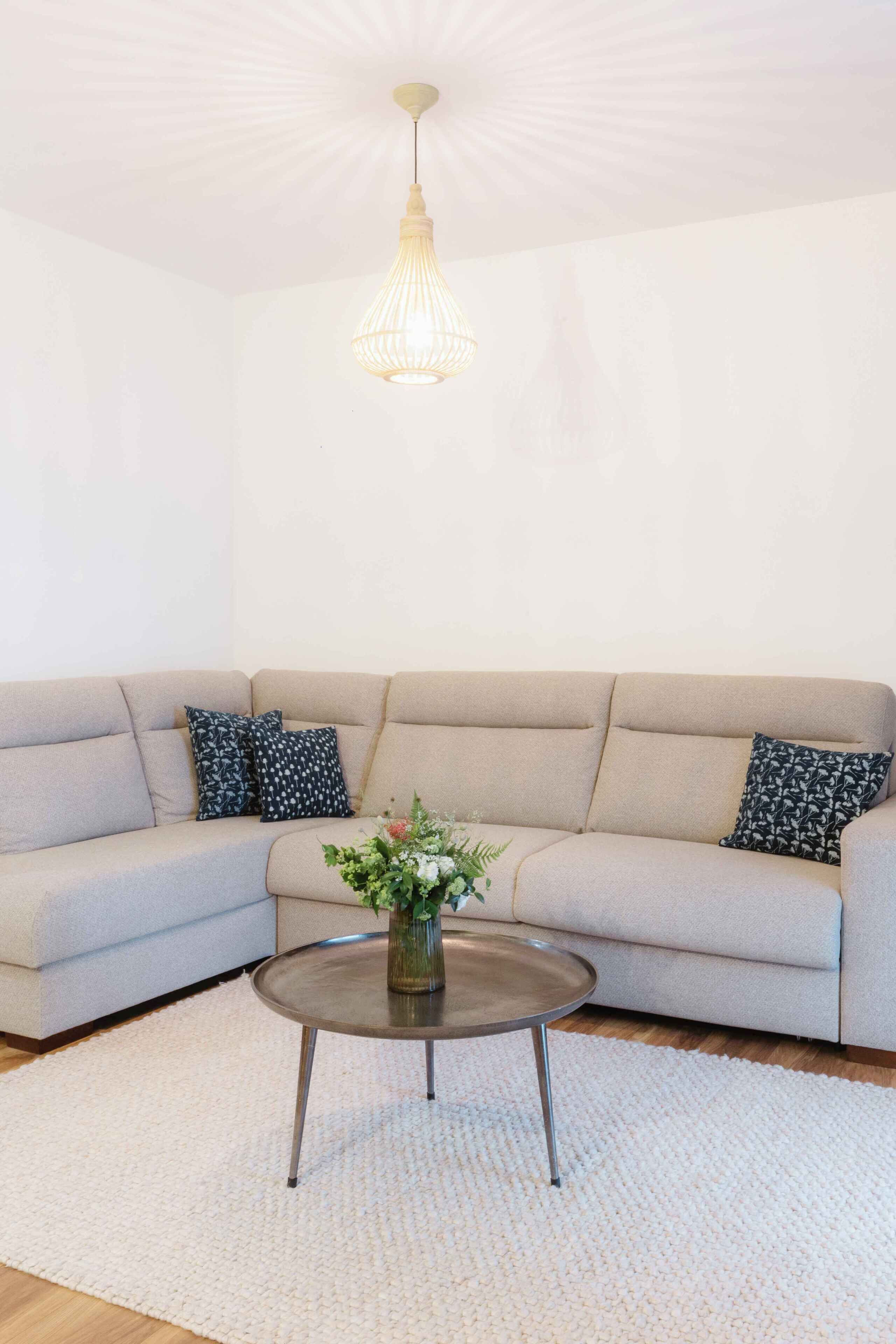 A living room with a beige sofa, two patterned pillows, a glass coffee table with a vase of flowers, and a pendant light.