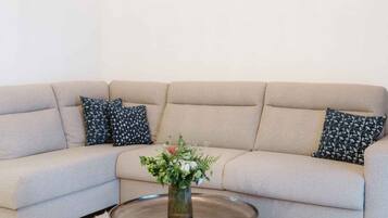 A living room with a beige sofa, two patterned pillows, a glass coffee table with a vase of flowers, and a pendant light.