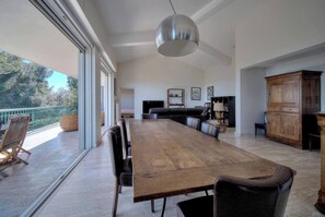A spacious dining area with a long wooden table and chairs, a large pendant light, and a view of the outdoors through large windows.
