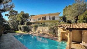 A swimming pool with a wooden deck, a stone wall, and a two-story building with balconies. - FRGK Villa Le Petit Bois Panoramic View, Lush Ga (vallauris)