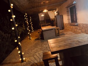 An outdoor patio area with stone flooring, wooden tables and benches, string lights, and a stone fireplace. - Beautiful B&B & Villa Casa Huerta Del Rio (Riogordo)