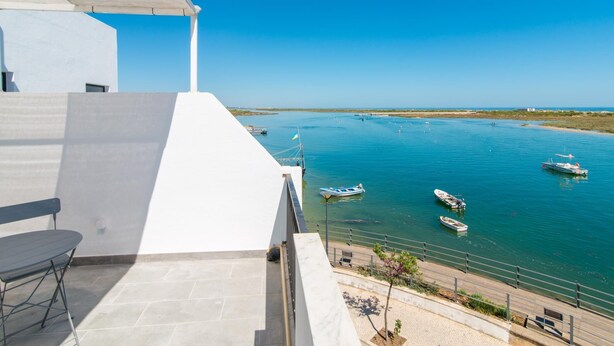A terrace with a view of a calm bay with several boats, a white building, and a clear blue sky. - Studio Apartment with Sea View (Cabanas de Tavira)