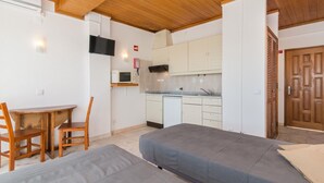 A compact room with a wooden ceiling, a kitchenette, a TV mounted on the wall, and a bed with a grey mattress. - Studio Apartment with Sea View (Cabanas de Tavira)
