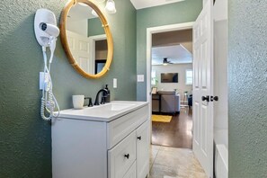 A bathroom with a white vanity, a round mirror, and a hairdryer on the wall. - The Cactus Rose - 2BR 1BA - Walk to Texas Tech (Lubbock)