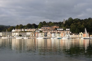 A waterfront property with a row of buildings, a marina with boats, and a hill with trees in the background.