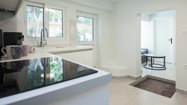 A modern kitchen with a large island, white cabinetry, and a view of the outdoors through large windows. - Cozy 1 Bd Basement Apt in Pagrati (Athens)