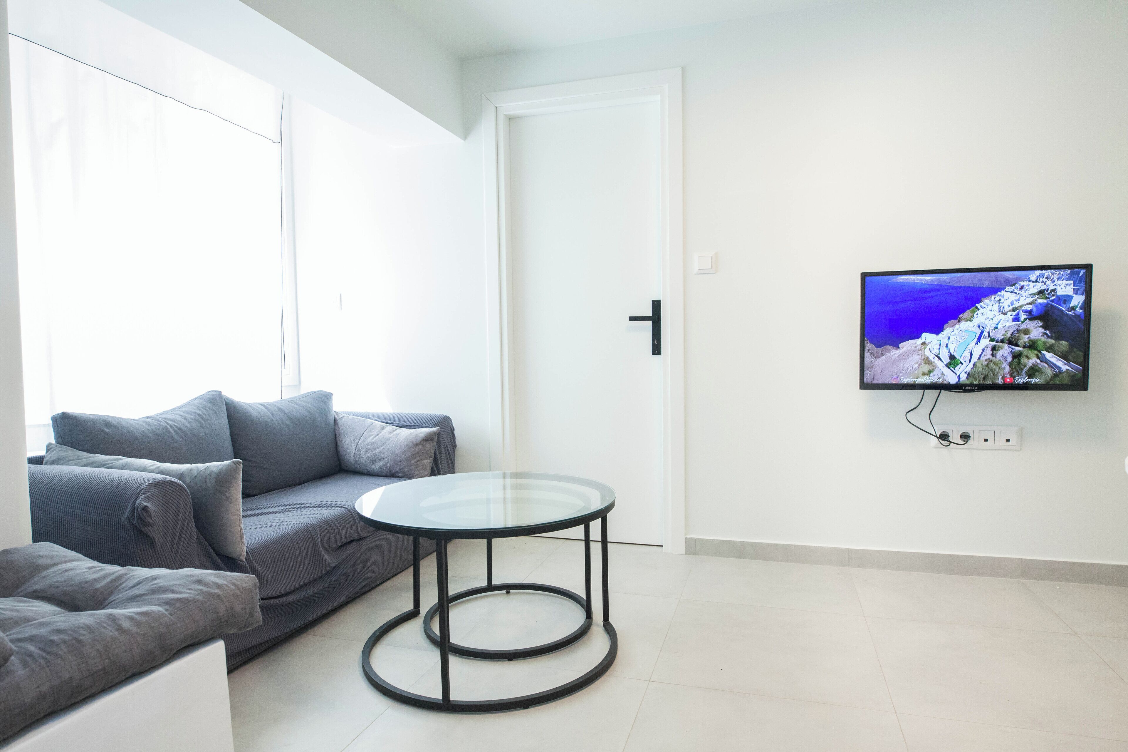 A modern living room with a grey sofa, a glass-top coffee table, and a flat-screen TV mounted on the wall.
