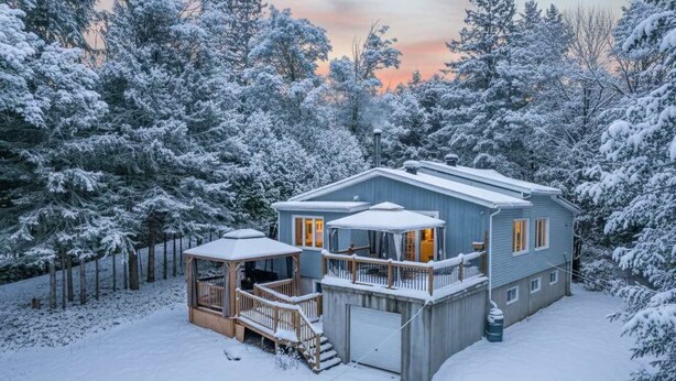 A two-story house with a covered porch and a garage, surrounded by snow-covered trees. - Spa & Nature | Le Havre du Confort (Saint-Colomban)