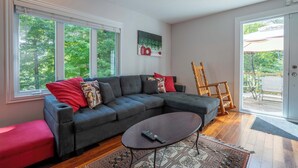 A modern living room with a grey sectional sofa, a wooden coffee table, and a large window offering a view of trees. - Spa & Nature | Le Havre du Confort (Saint-Colomban)