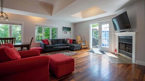 A spacious living room with a dark-colored sectional sofa, a red armchair, and a flat-screen TV mounted above a fireplace. - Spa & Nature | Le Havre du Confort (Saint-Colomban)