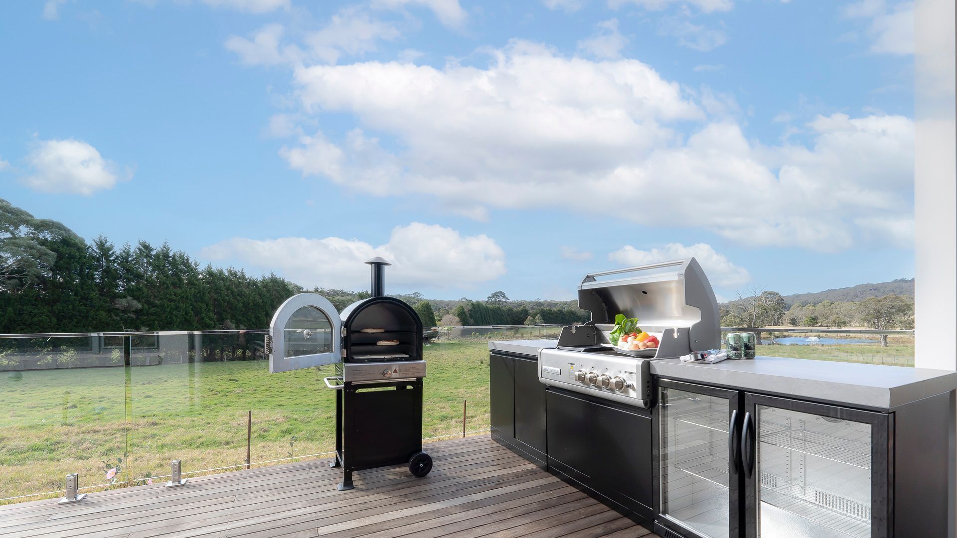 Cocina exterior con parrilla, horno y barra en una terraza con vistas a un campo cubierto de hierba.