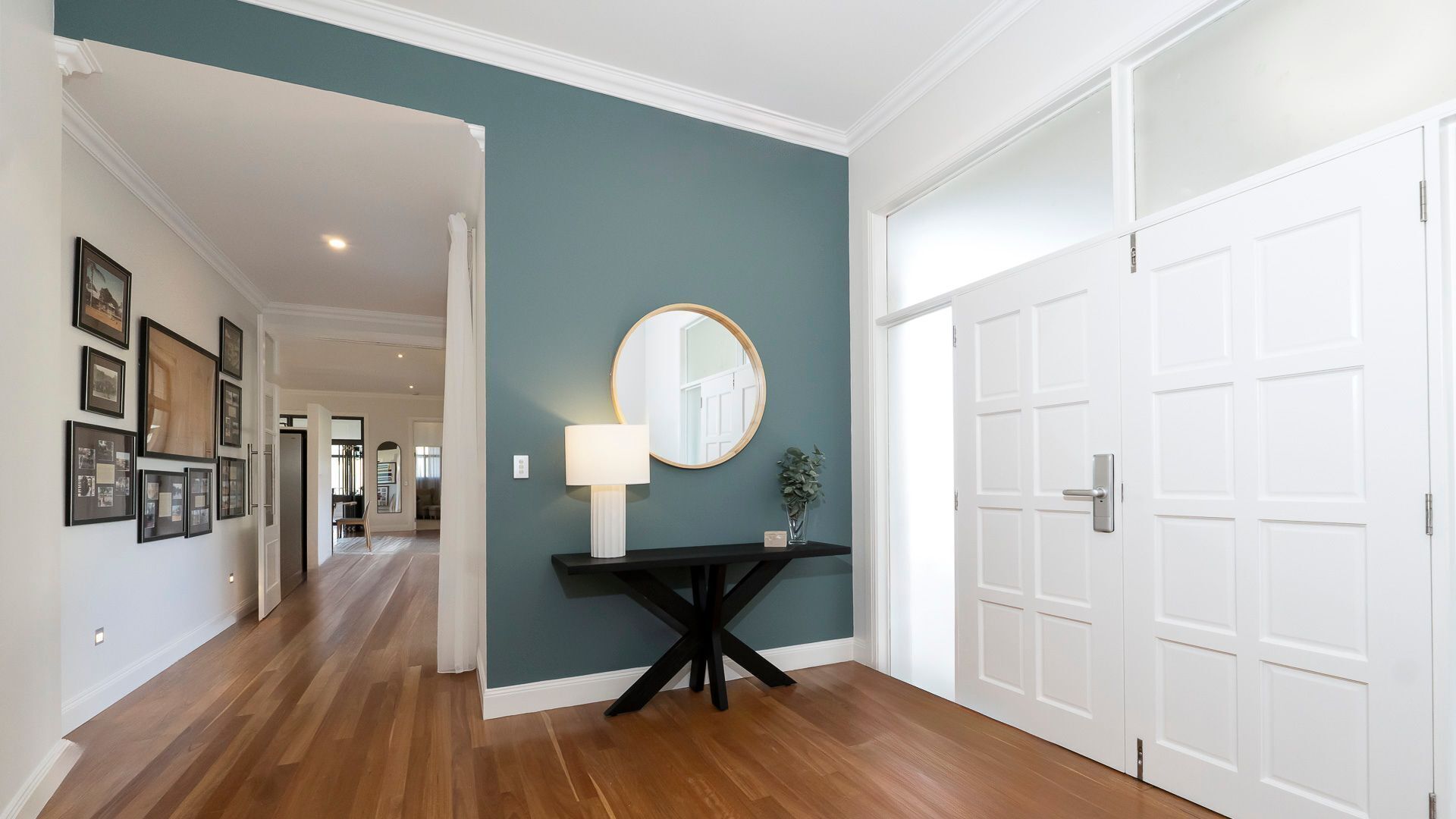 A hallway with a wooden floor, a black console table with a lamp and mirror, and white double doors.
