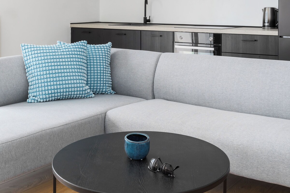 A modern living room with a grey sofa, a round black coffee table, and a kitchen area with dark cabinets and a white countertop.