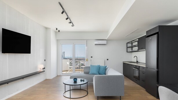 Um apartamento moderno com sala de estar, sofá cinzento, mesa de centro, televisão de tela plana, cozinha com eletrodomésticos pretos e varanda com vista para a cidade.