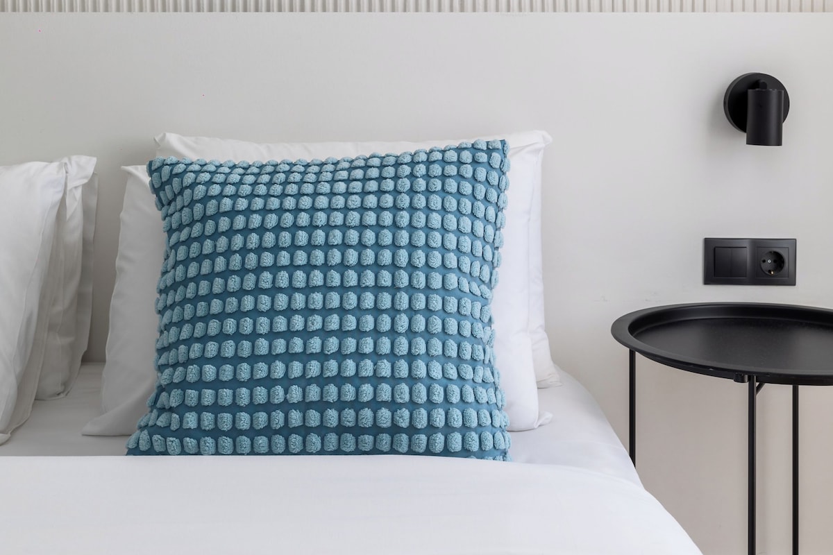 A neatly made bed with white and blue patterned pillows, a black side table, and a wall-mounted light fixture.