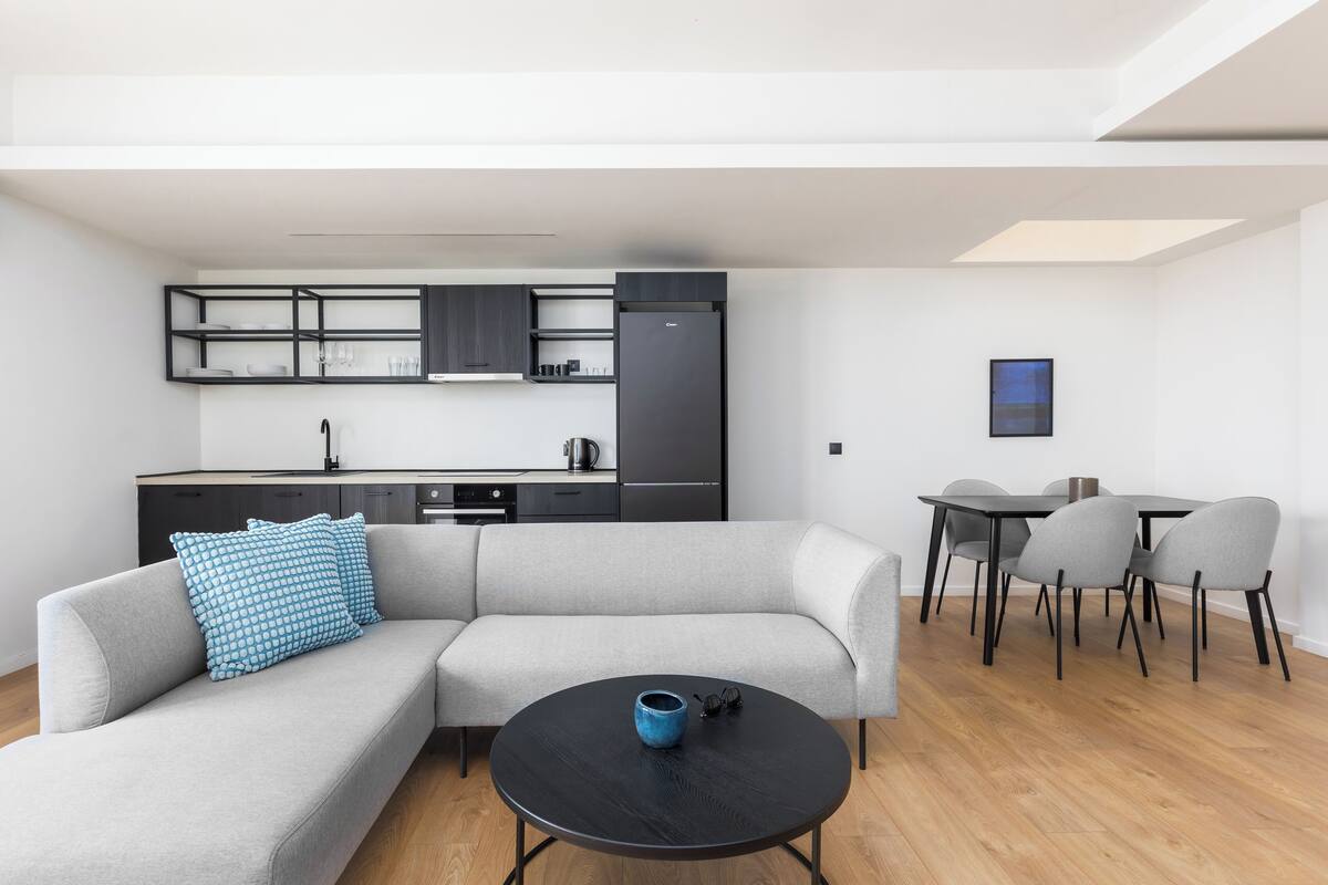 A modern living space with a sofa, a round coffee table, a dining table with chairs, and a kitchen area with cabinets and appliances.