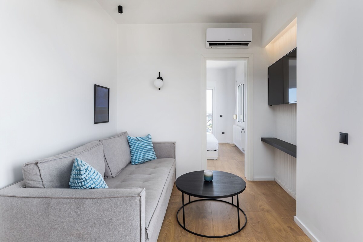 A modern living room with a grey sofa, a round black coffee table, and a flat-screen TV mounted on the wall.