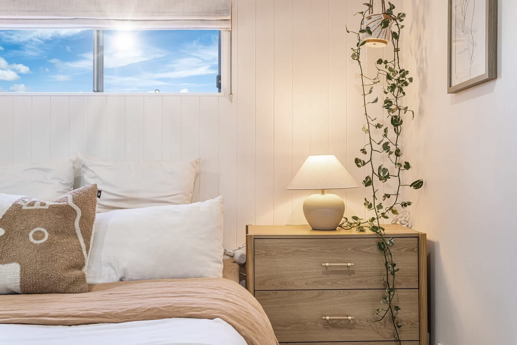 Un dormitorio con cama, mesita de noche con lámpara, un tocador de madera y ventana con vista al cielo.